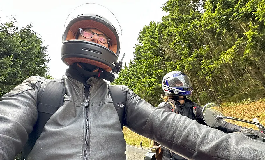 Zwei Motorradfahrer:innen mit Sena Intercom-Systemen am Helm fahren auf einer Landstraße durch ein bewaldetes Gebiet.