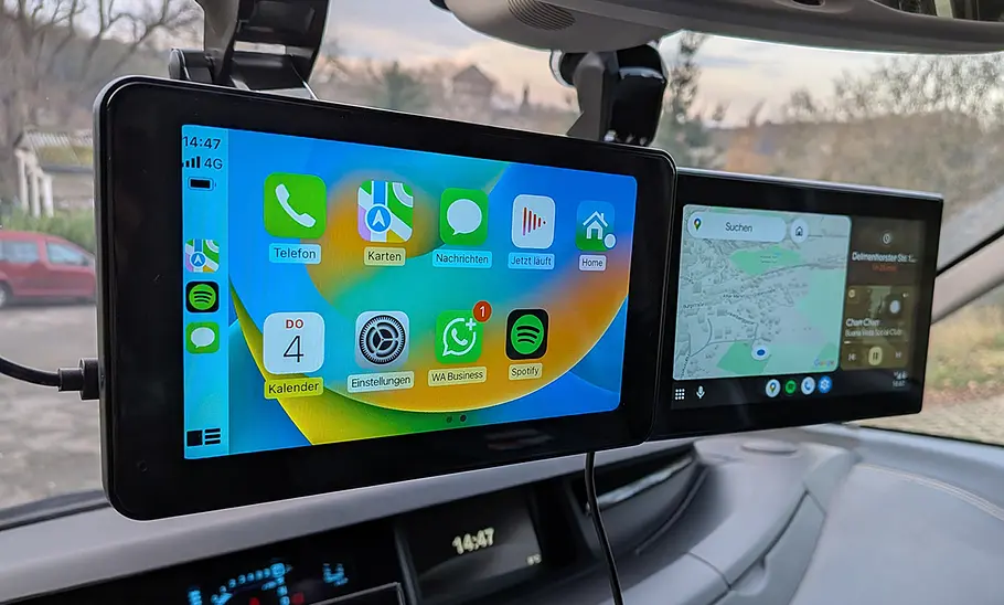 Zwei Android Auto und CarPlay-Displays im Testverfahren an einer Windschutzscheibe, fotografiert von schräg vorne.