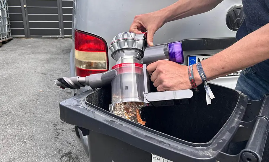 Die Außenaufnahme zeigt, wie der Auffangbehälter des Dyson Akku-Staubsauger Car & Boat V8 über einer Mülltonne geleert wird.