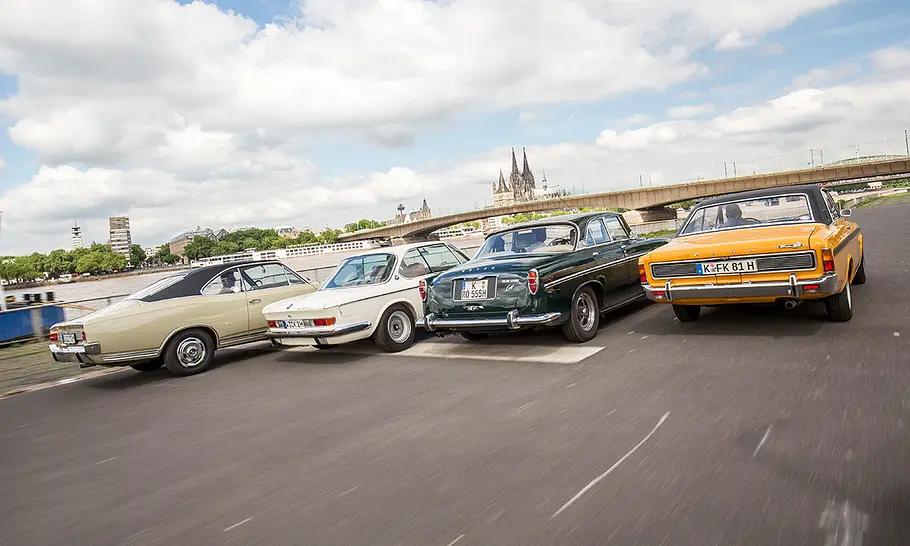 Die Autos Opel Commodore A Coupé, BMW 2800 CS, Rover P5B Coupé & Ford 20M RS Coupé fahren an einer Straße am Rhein entlang, fotografiert von schräg hinten.