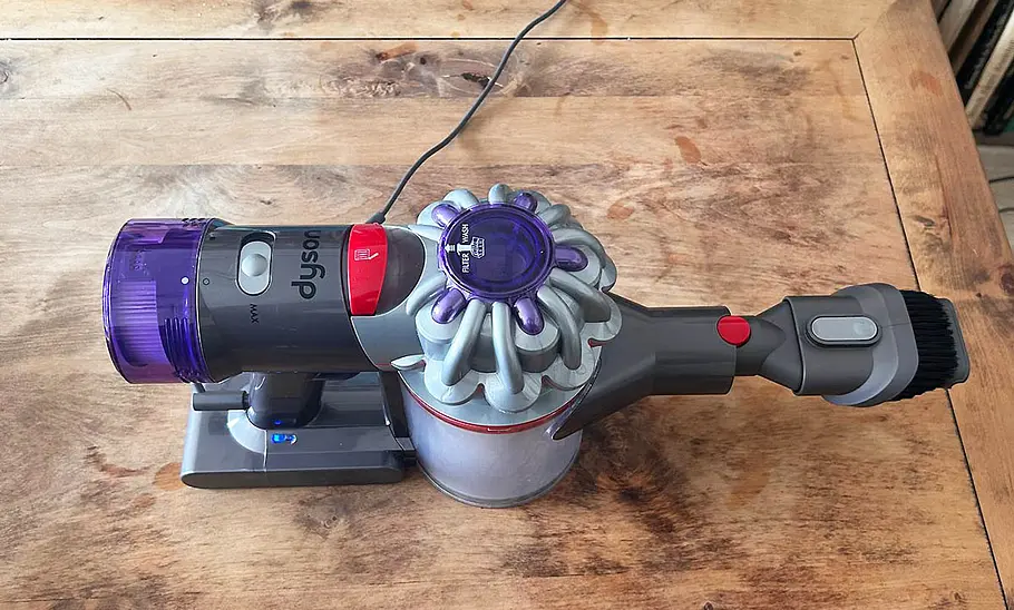Die Innenaufnahme zeigt, wie der Dyson Akku-Staubsauger Car & Boat V8 auf einem Holztisch liegt und per Kabel an einer Steckdose geladen wird.