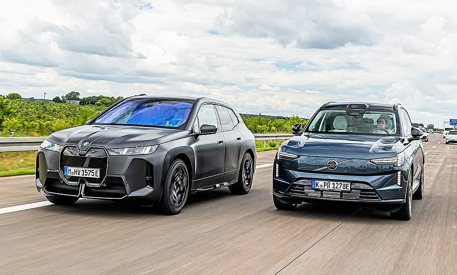 Blick von vorne auf die beiden Elektroautos BMW iX und Volvo EX90, die auf der Autobahn nebeneinander her fahren.