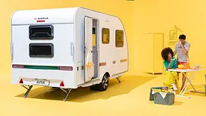 Der Adria Aviva Lite statsich von schräg hinten links in einem gelb gestrichenen Studio. Rechts vom Caravan steht der herausnehmbare Tisch, an dem sich zwei Personen aufhalten. - Foto: Adria