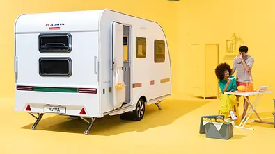 Der Adria Aviva Lite statsich von schräg hinten links in einem gelb gestrichenen Studio. Rechts vom Caravan steht der herausnehmbare Tisch, an dem sich zwei Personen aufhalten. - Foto: Adria