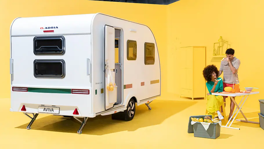 Der Adria Aviva Lite in einem gelben Studio, Ansicht von schräg hinten rechts. Am rechten Bildrand sitzen zwei Personen an einem Campingtisch.