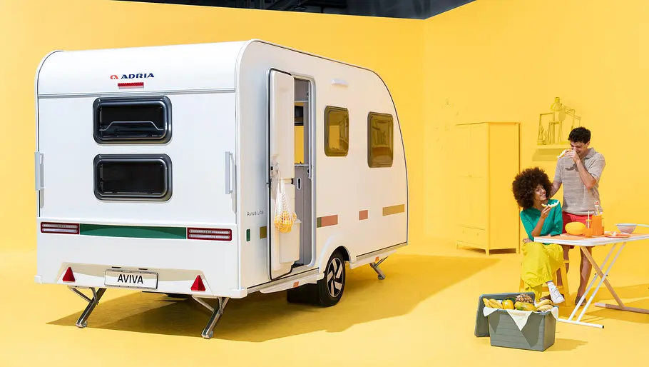 Adria Aviva Lite 360 DK Der Adria Aviva Lite statsich von schräg hinten links in einem gelb gestrichenen Studio. Rechts vom Caravan steht der herausnehmbare Tisch, an dem sich zwei Personen aufhalten.