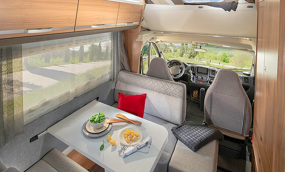 Das Bild zeigt die Sitzgruppe des Adria Coral XL 600 DP (2024) mit Blick in Richtung Cockpit. Das Alkovenbett ist hochgefahren.