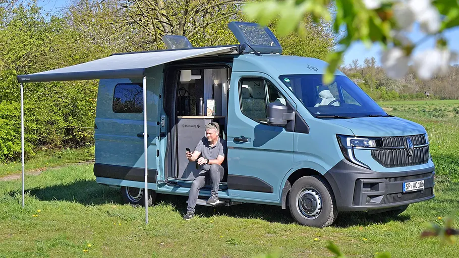 Der Ahorn Camp CV 560 statisch von schräg vorne rechts mit geöffneter Markise auf einer Wiese stehend. Redakteur Martin Urbanke sitzt im Einstiegsbereich der geöffneten Schiebetüre.