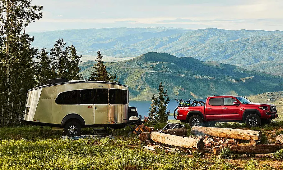Airstream Basecamp 20 (2025) Das Bild zeigt den Airstream Basecamp 20 (2025) stehend von schräg rechts fotografiert. Im Hintergrund ist ein Bergpanorama zu sehen, vor dem Caravan steht ein roter Toyota Tacoma mit Fahrrädern auf der Ladefläche.