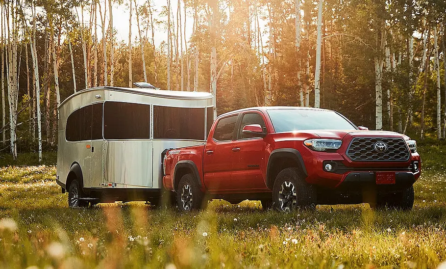 Airstream Basecamp 20 (2025) Das Bild zeigt den Airstream Basecamp 20 (2025) stehend von schräg vorne rechts fotografiert. Der Caravan hängt an einem roten Toyota Tacoma. Im Hintergrund ist ein Wald zu sehen.
