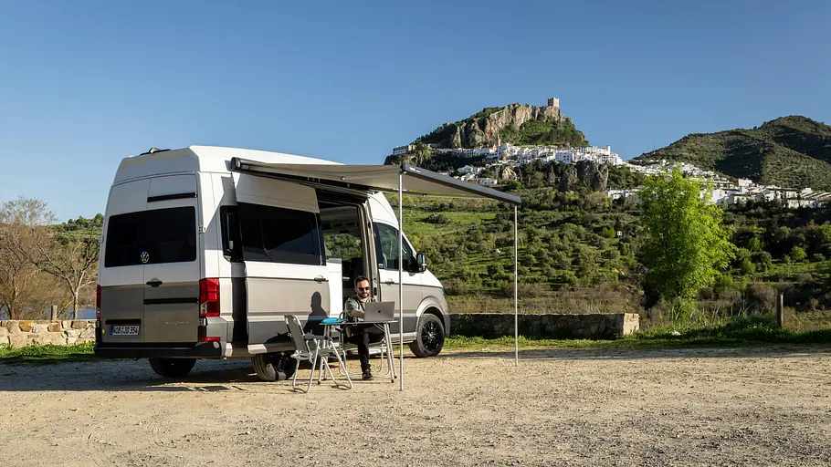 Der VW Grand California mit ausgefahrener Markise von schräg hinten rechts. Unter der Markise sitzt Autor Dominik Mothes auf Campingmöbeln, im Hintergrund ist die Stadt Zahara de la Sierra zu sehen.