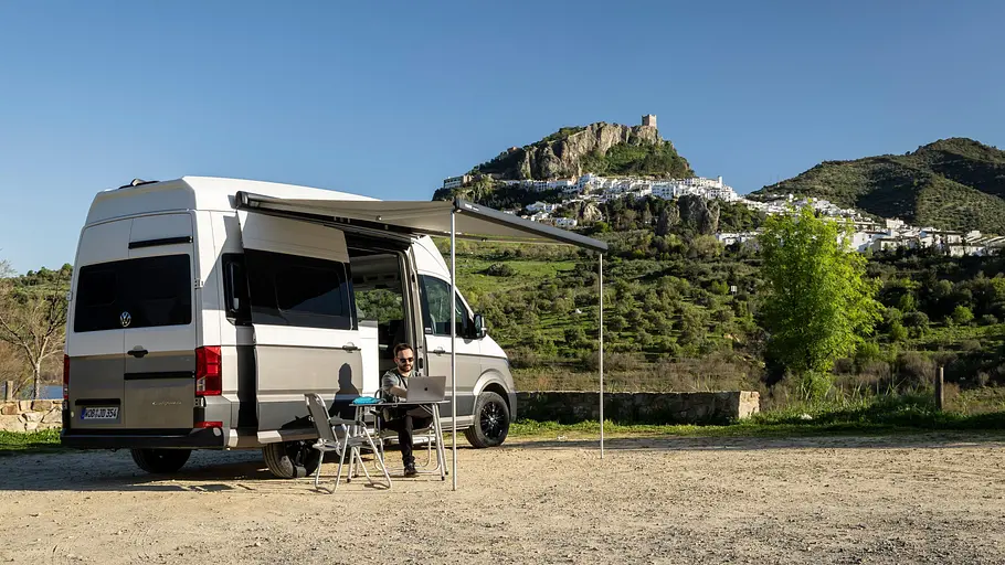 Der VW Grand California mit ausgefahrener Markise von schräg hinten rechts. Unter der Markise sitzt Autor Dominik Mothes auf Campingmöbeln, im Hintergrund ist die Stadt Zahara de la Sierra zu sehen.