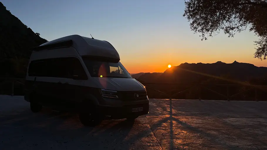 Der VW Grand California im Schatten statsich von schräg vorne rechts mit Sonnenuntergang im Hintergrund.
