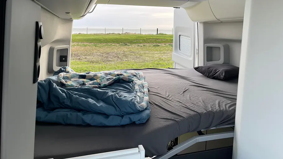 Das Heckbett im VW Grand California 600 mit Blick aufs Meer durch die geöffneten Hecktüren.