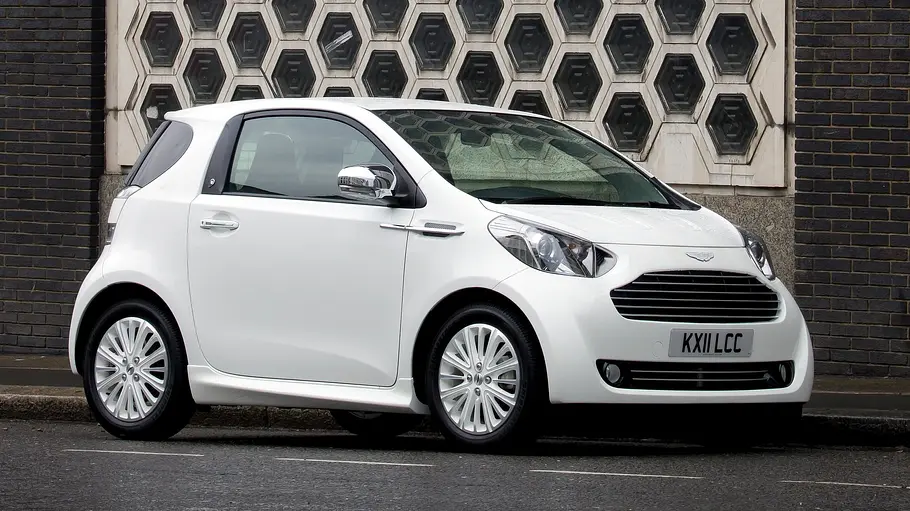 Der Aston Martin Cygnet steht vor einer Ziegelsteinfassade, von vorne rechts fotografiert.