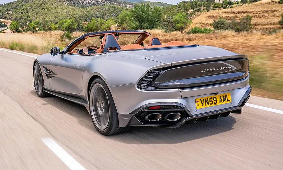 Der Aston Martin Vanquish Volante fahrend von schräg hinten mit offenem Verdeck. Redakteurin Adele Moser am Steuer.