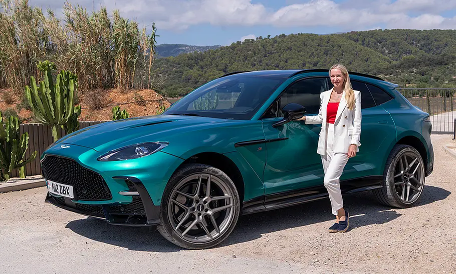 Der Aston Martin DBX S (2025) stehend von der Seite, daneben steht Redakteurin Adele Moser.