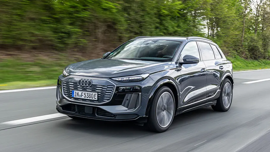 Ein grauer Audi Q6 e-tron fährt auf einer Straße an grünen Bäumen vorbei. Das Fahrzeug ist schräg von vorn zu sehen.