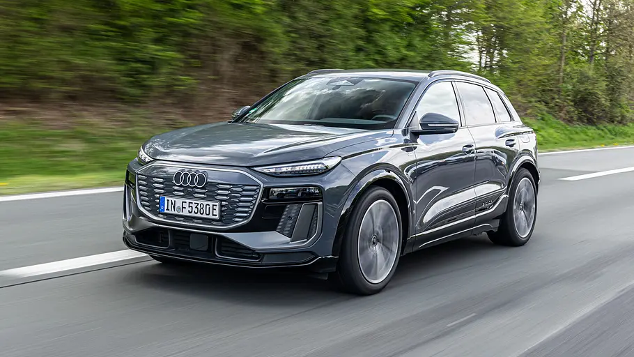 Ein grauer Audi Q6 e-tron quattro fährt auf einer Straße an grünen Bäumen vorbei. Das Fahrzeug ist schräg von vorn zu sehen.