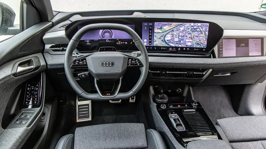 Das Audi Q6 e-tron Cockpit zeigt ein Lenkrad, digitale Anzeigen mit Navigationskarte, einen Beifahrerbildschirm und graue Stoffsitze.