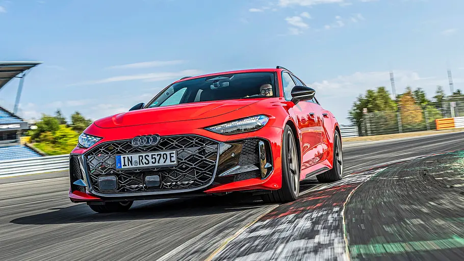 Audi RS 5 Avant Tracktest Ein roter Audi RS 5 Avant fährt durch eine Kurve auf einer Rennstrecke. Im Hintergrund sind Tribünen und blauer Himmel zu sehen.
