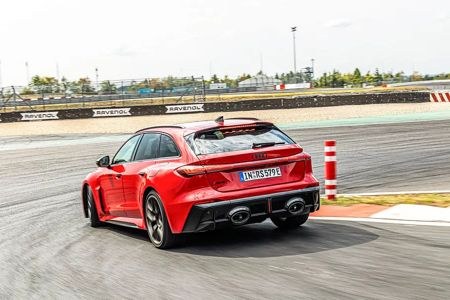 Audi RS 5 Avant im Tracktest Ein roter Audi RS 5 Avant fährt durch eine Kurve auf einer Rennstrecke, von schräg hinten neben rot-weißen Randmarkierungen gesehen.