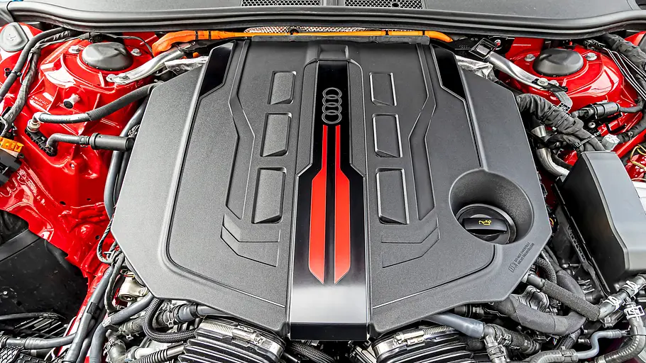 Audi RS 5 Avant Tracktest Im roten Motorraum sitzt eine schwarze Motorabdeckung mit Audi-Ringen und roten Streifen, umgeben von Schläuchen und Kabeln.
