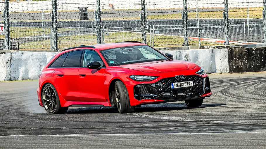 Audi RS 5 Avant Tracktest Ein roter Audi RS 5 Avant driftet durch eine Kurve auf einer Rennstrecke. Im Hintergrund sind Tribünen und blauer Himmel zu sehen.