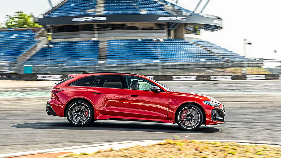Audi RS 5 Avant Tracktest Ein roter Audi RS 5 Avant fährt durch eine Kurve auf einer Rennstrecke. Im Hintergrund sind Tribünen und blauer Himmel zu sehen.