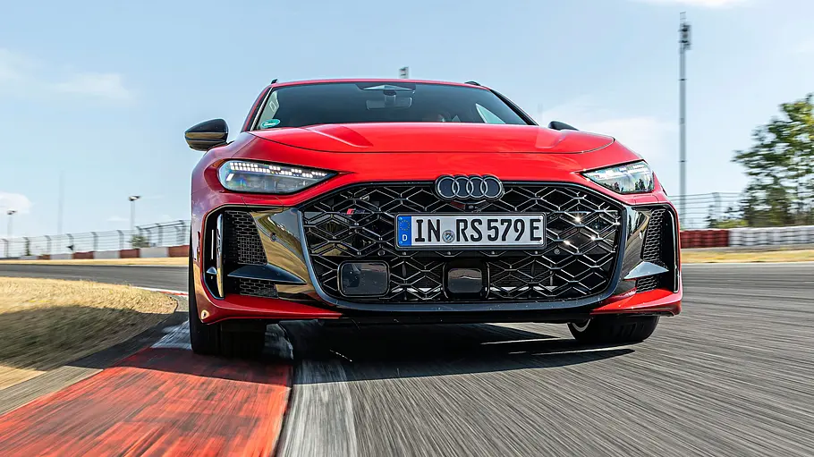 Audi RS 5 Avant Tracktest Ein roter Audi RS 5 Avant fährt durch eine Kurve auf einer Rennstrecke. Im Hintergrund sind Tribünen und blauer Himmel zu sehen.