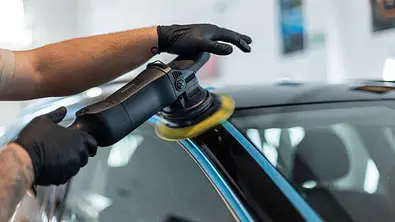 Eine Person poliert ein schwarz lackiertes Auto an der A-Säule mit einer Poliermaschine. Kunststoff- und Chromteile sind zum Schutz mit blauem Klebeband abgeklebt. - Foto: Imago