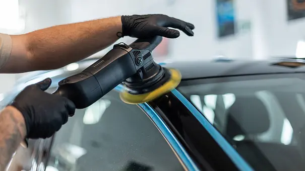 Eine Person poliert ein schwarz lackiertes Auto an der A-Säule mit einer Poliermaschine. Kunststoff- und Chromteile sind zum Schutz mit blauem Klebeband abgeklebt. - Foto: Imago Eine Person poliert ein schwarz lackiertes Auto an der A-Säule mit einer Poliermaschine. Kunststoff- und Chromteile sind zum Schutz mit blauem Klebeband abgeklebt. - Foto: Imago