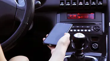 Eine Detailaufnahme von einer Hand, die ein Handy festhält. Im Hintergrund sieht man das Armaturenbrett eines Autos. - Foto: iStock/triocean