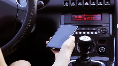 Eine Detailaufnahme von einer Hand, die ein Handy festhält. Im Hintergrund sieht man das Armaturenbrett eines Autos. - Foto: iStock/triocean