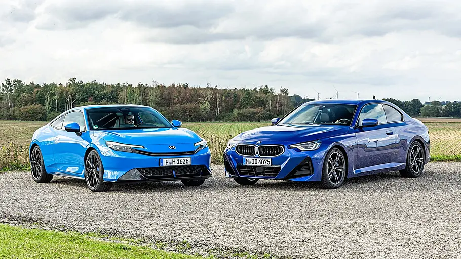 BMW 2er Coupé gegen Honda Prelude im Vergleichstest Ein hellblauer Honda Prelude und ein dunkelblaues BMW 220 Coupé stehen auf einem Schotterweg vor Feldern unter einem bewölkten Himmel.