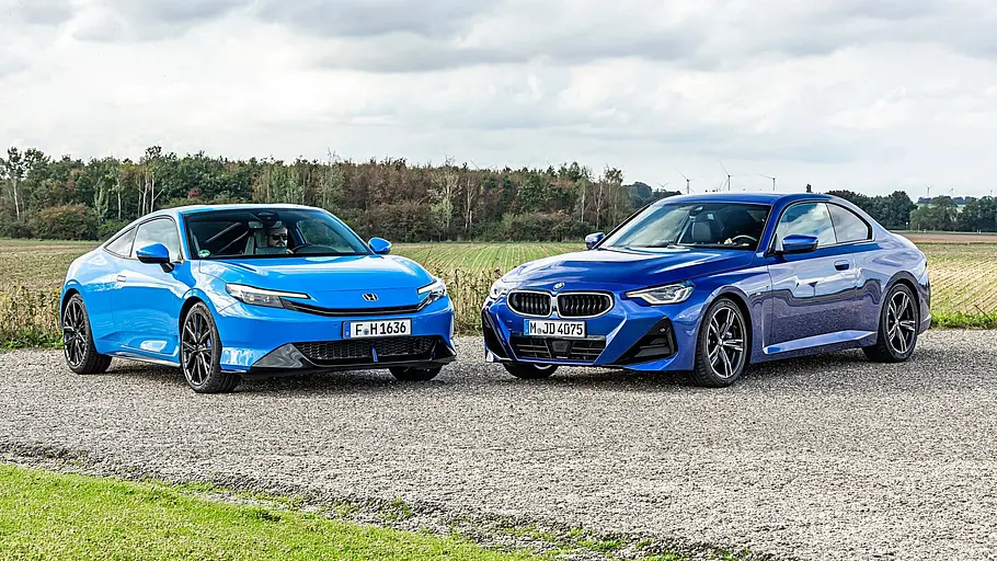 BMW 2er Coupé gegen Honda Prelude im Vergleichstest Ein hellblauer Honda Prelude und ein dunkelblauer BMW 220i M Sport Coupé stehen auf einem Schotterweg vor Feldern unter einem wolkigen Himmel.