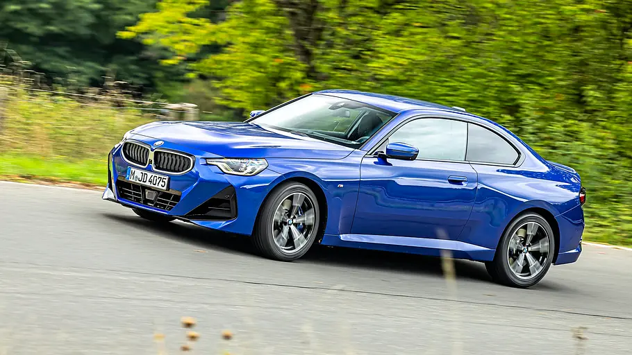 BMW 2er Coupé gegen Honda Prelude im Vergleichstest Ein blaues BMW 220i M Sport Coupé fährt auf einer Straße an grünen Bäumen vorbei, schräg von vorn und von der Seite aufgenommen.