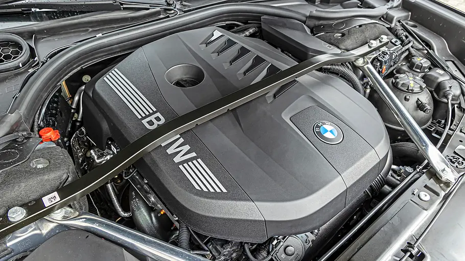 BMW 740 xDrive Einzeltest 7er Facelift Im offenen Motorraum des BMW 740 xDrive sind eine schwarze Motorabdeckung mit Logo, Metallstreben, Schläuche und Behälter zu sehen.
