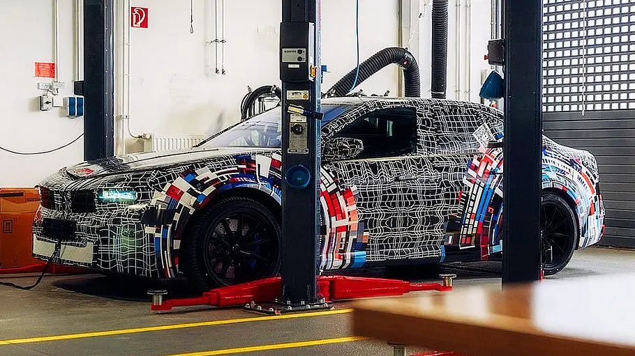 BMW i3 M (2027) Blick von schräg vorne auf den BMW i3 M (2027), der in einer Werkstatthalle zwischen den Säulen einer Hebebühne steht.