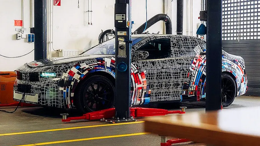 Blick von schräg vorne auf den BMW i3 M (2027), der in einer Werkstatthalle zwischen den Säulen einer Hebebühne steht.