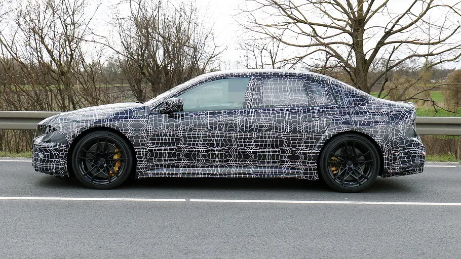 BMW i3 M (2027) Der BMW i3 M fährt als Erlkönig auf einer Landstraße, von links fotografiert.