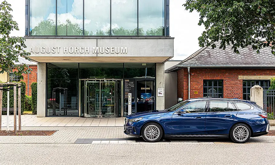 BMW i5 Touring Blick von der Seite auf den BMW i5 Touring, der neben einem Gebäude am Straßenrand parkt.