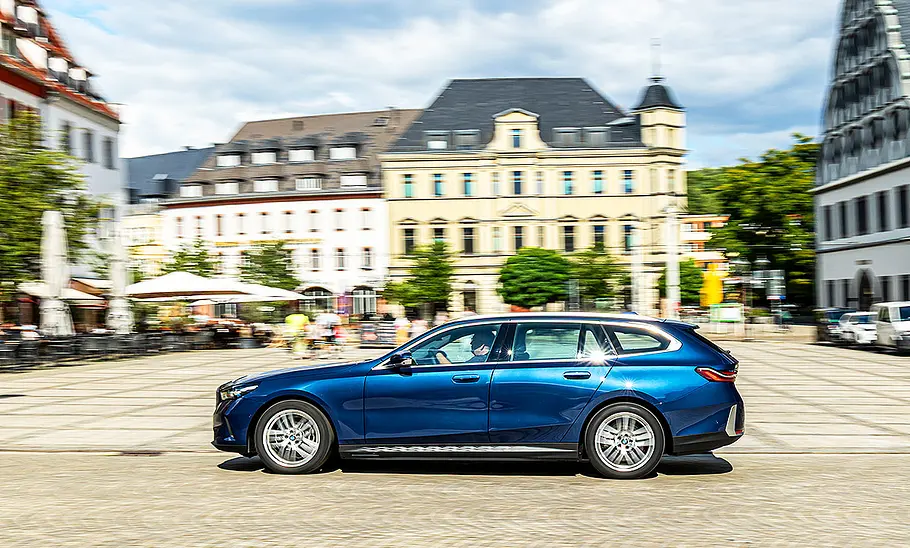 BMW i5 Touring Blick von der Seite auf den fahrenden BMW i5 Touring, der durch eine Innenstadt an einem Marktplatz entlang fährt.