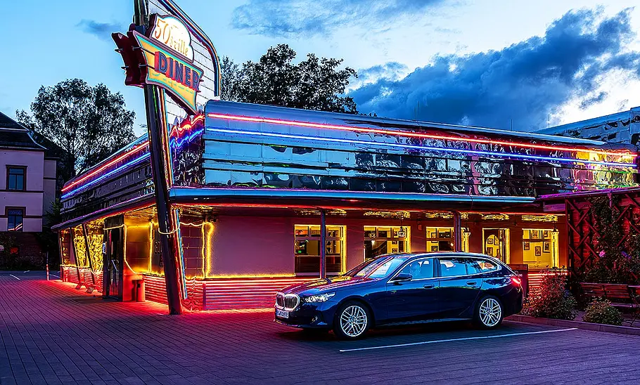 BMW i5 Touring Blick von schräg vorne auf den BMW i5 Touring, der abends vor einem beleuchteten Diner parkt.