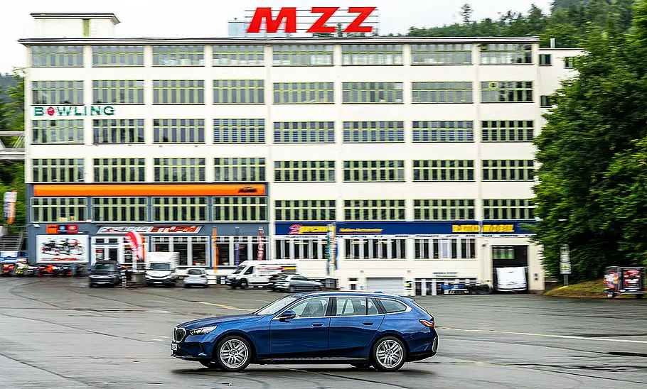 BMW i5 Touring Blick von der Seite auf den fahrenden BMW i5 Touring, der vor einem großen Gebäude entlang fährt.