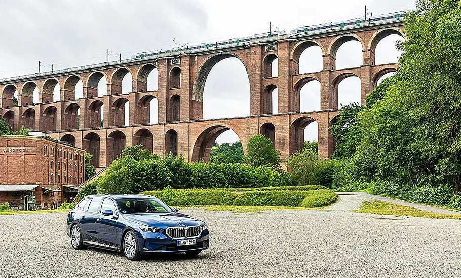 BMW i5 Touring Blich von schräg vorne auf den BMW i5 Touring, der vor einer großen Bogenbrücke posiert.