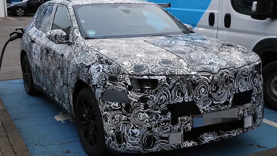Als Erlkönig erwischt: Der BMW iX1 steht an einer Ladesäule, fotografiert von schräg vorne.
