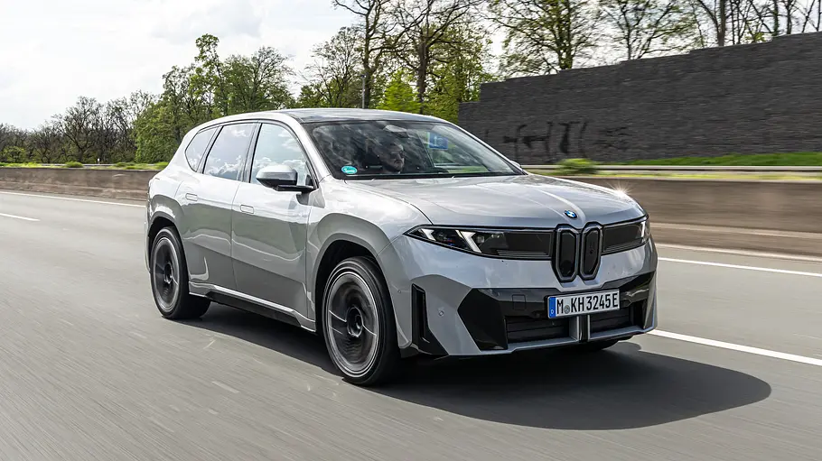 Ein silberner BMW iX3 50 xDrive fährt auf einer mehrspurigen Straße. Im Hintergrund stehen Bäume hinter einer Lärmschutzwand.