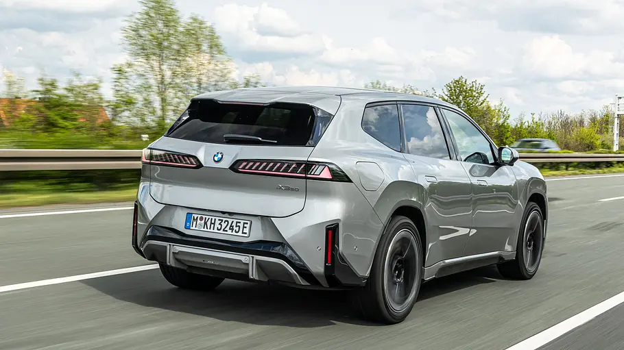 Ein silberner BMW iX3 fährt auf einer Autobahn, schräg von hinten gesehen. Im Hintergrund stehen Bäume unter bewölktem Himmel.