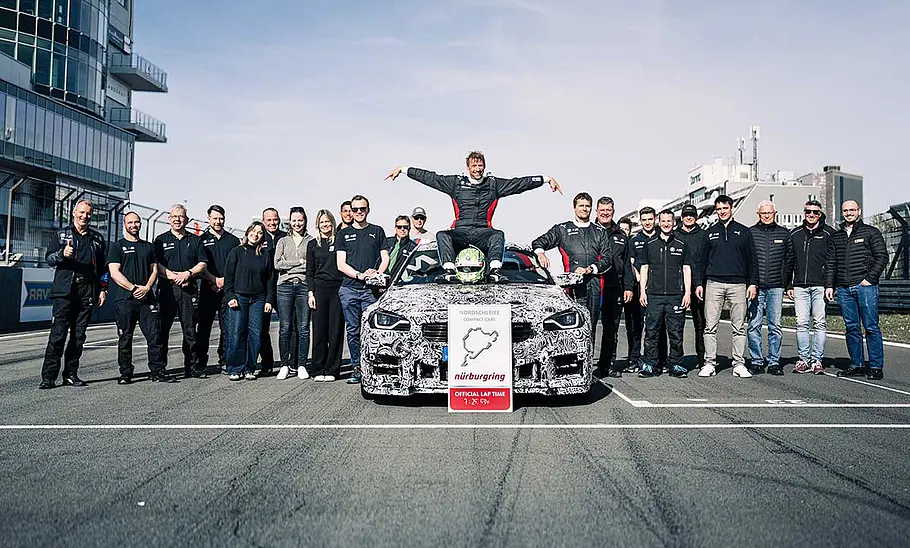 BMW M2 CS (2025) fährt Nordschleifen-Rekord: Bild zeigt Fahrer und Entwicklungsingenieur Jörg Weidinger auf der Motorhaube, daneben steht das Team, das ihn unterstützt hat.